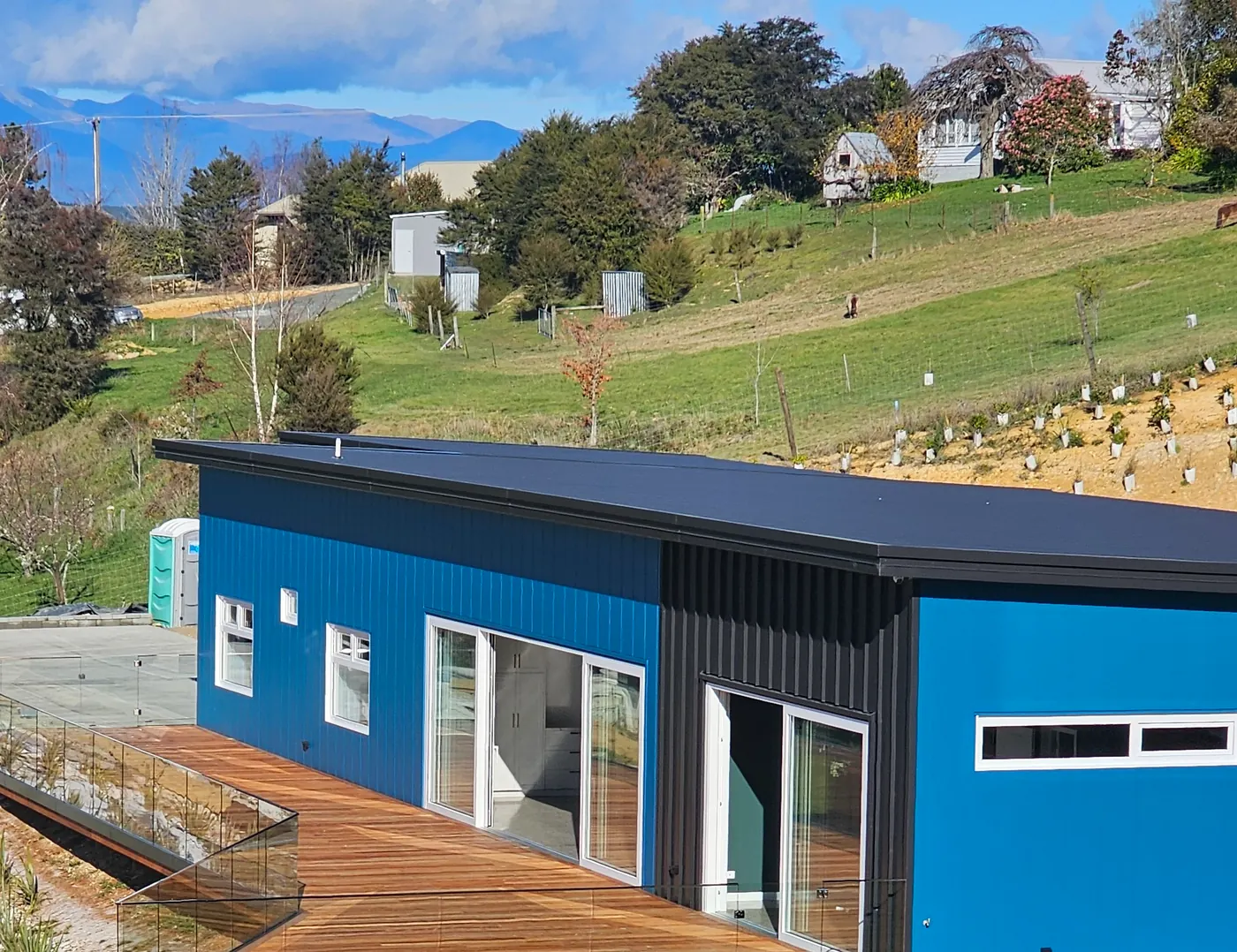 Blue-clad Ecotectural home with a timber deck and glass balustrade, looking out to a rural mountain outlook