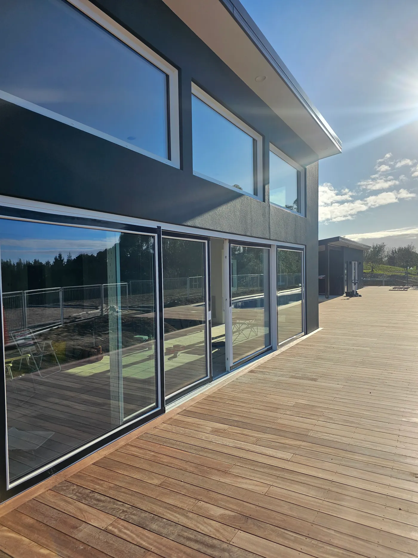 Dark-clad two-storey Ecotectural home with full-height glass sliders opening onto a sunlit timber deck and pool