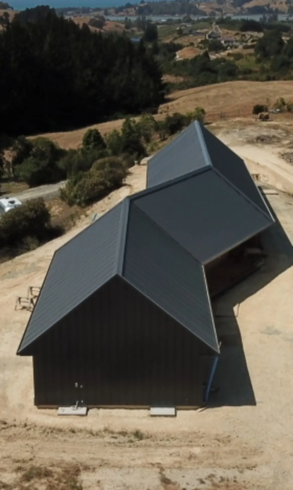 Aerial view of a dark-clad Ecotectural home on a rural hillside site, with the coast beyond