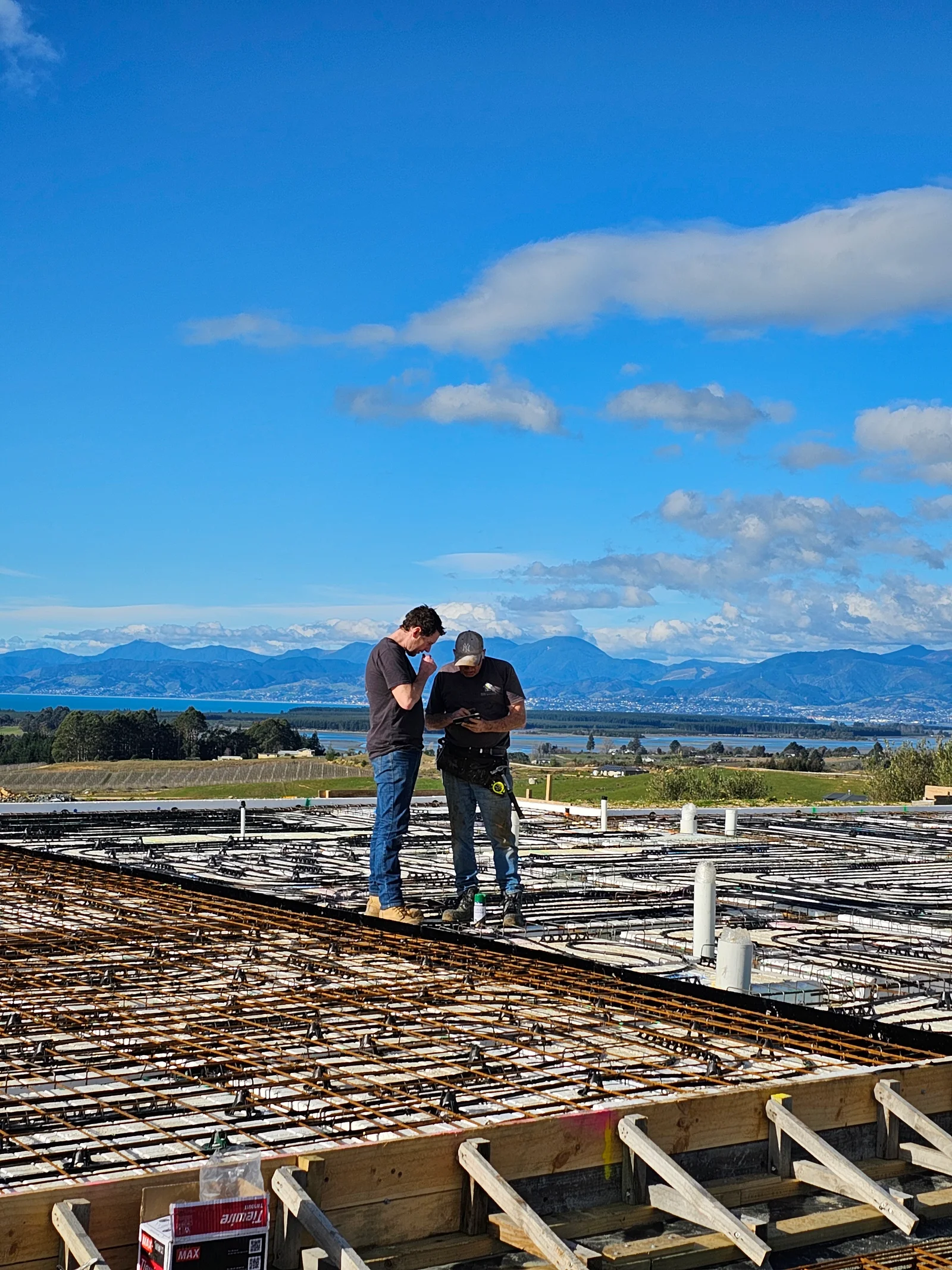 Daryl Combes reviewing the MAXRaft foundation slab and underfloor heating layout on a rural Tasman site, with Tasman Bay and the mountains beyond