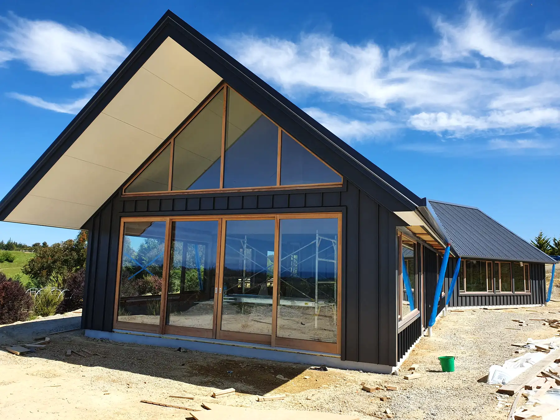 Dark-clad gable home under construction with full-height timber-framed glazing under a blue Tasman sky